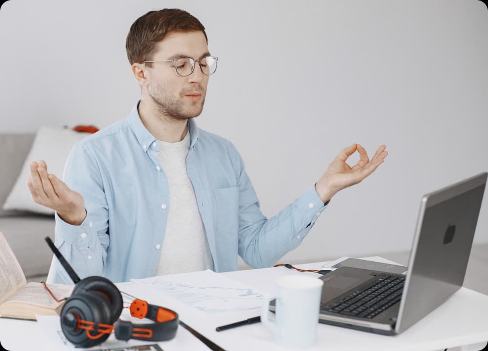 Man practicing mindfulness meditation at his desk while working on a laptop, promoting cognitive resilience in a high-pressure work setting.