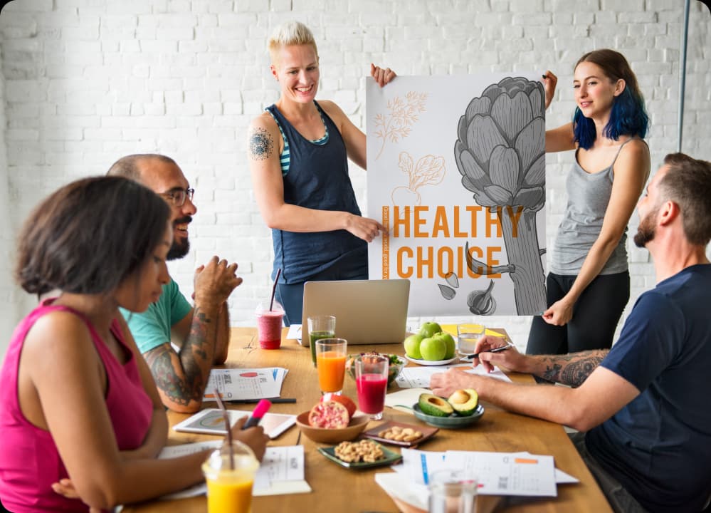 Team discussing healthy lifestyle choices with fresh food and a "Healthy Choice" poster, representing holistic health as a preventative strategy.