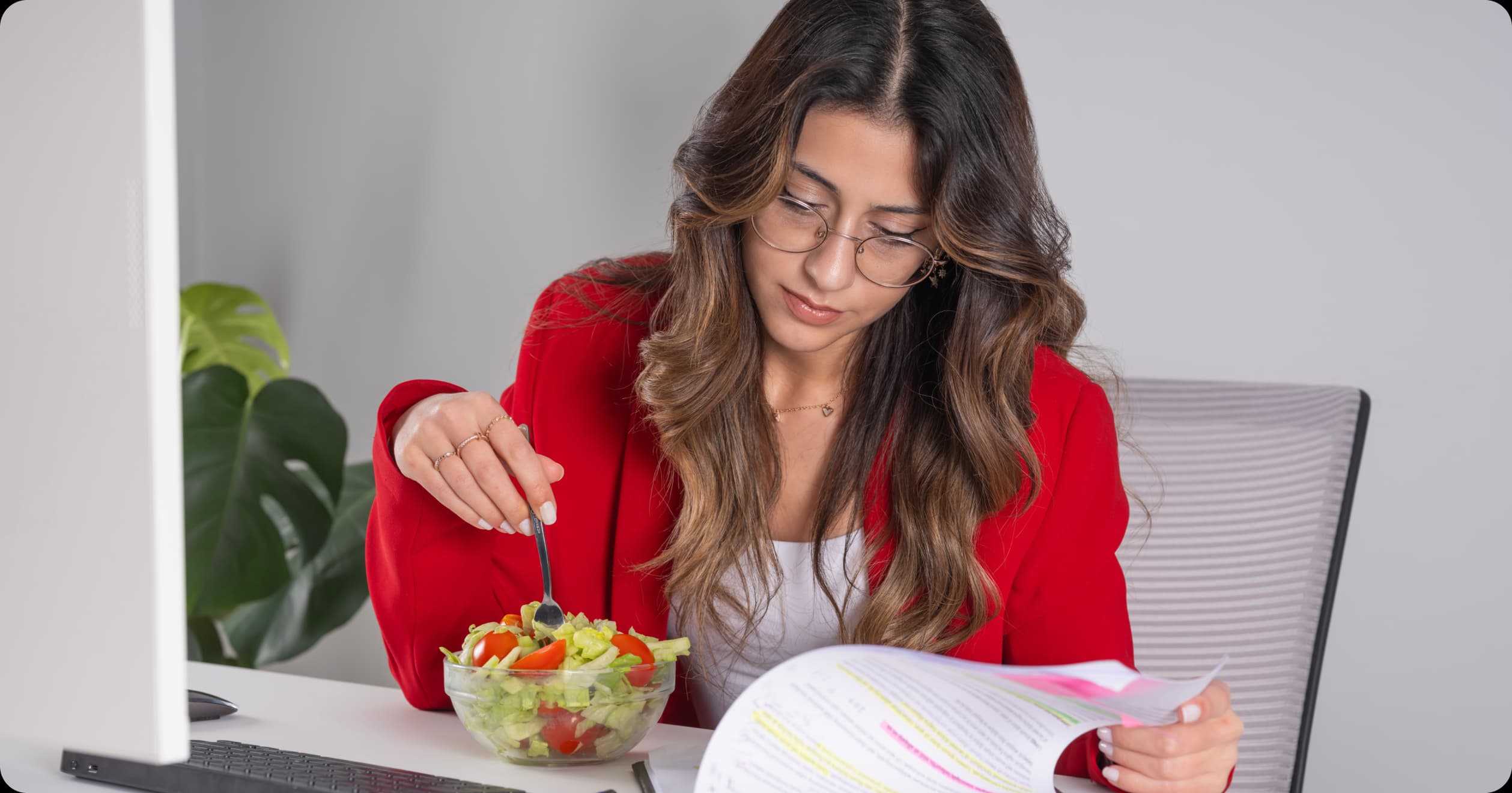 Woman eating a healthy salad while reviewing work documents at her desk, representing personalized nutrition as a key part of employee wellness.